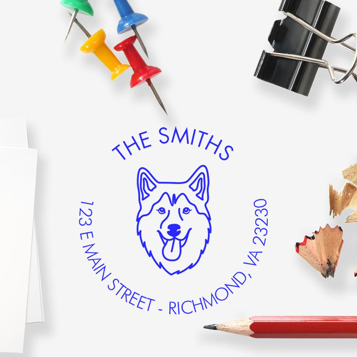 Blue custom dog address stamp reading The Smiths, with colorful push pins, black binder clip, and red pencil.