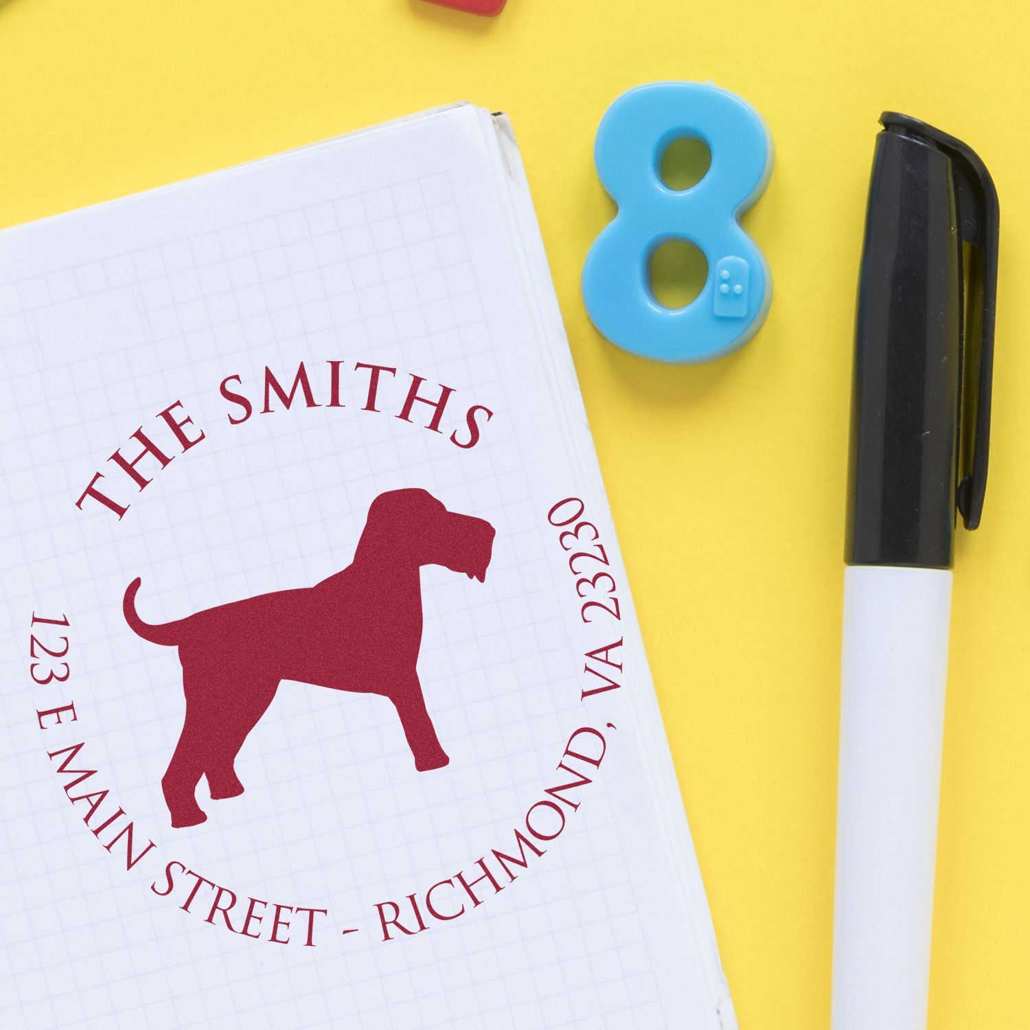 Red dog address seal stamp on notebook, with blue number 8 stamp and black pen on a bright yellow background.