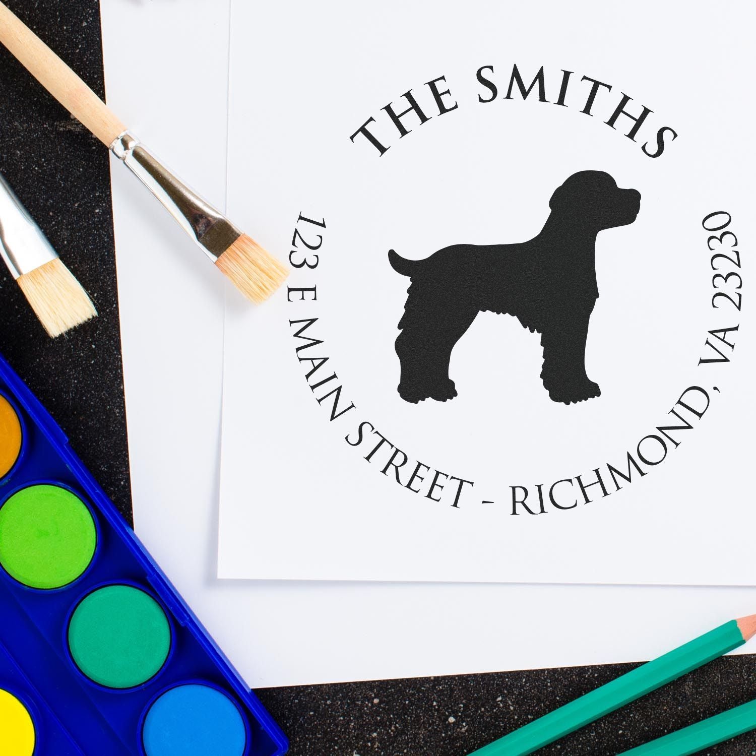 Custom black dog stamp seal for The Smiths on white paper, with paintbrushes, watercolor palette, and teal pencils.