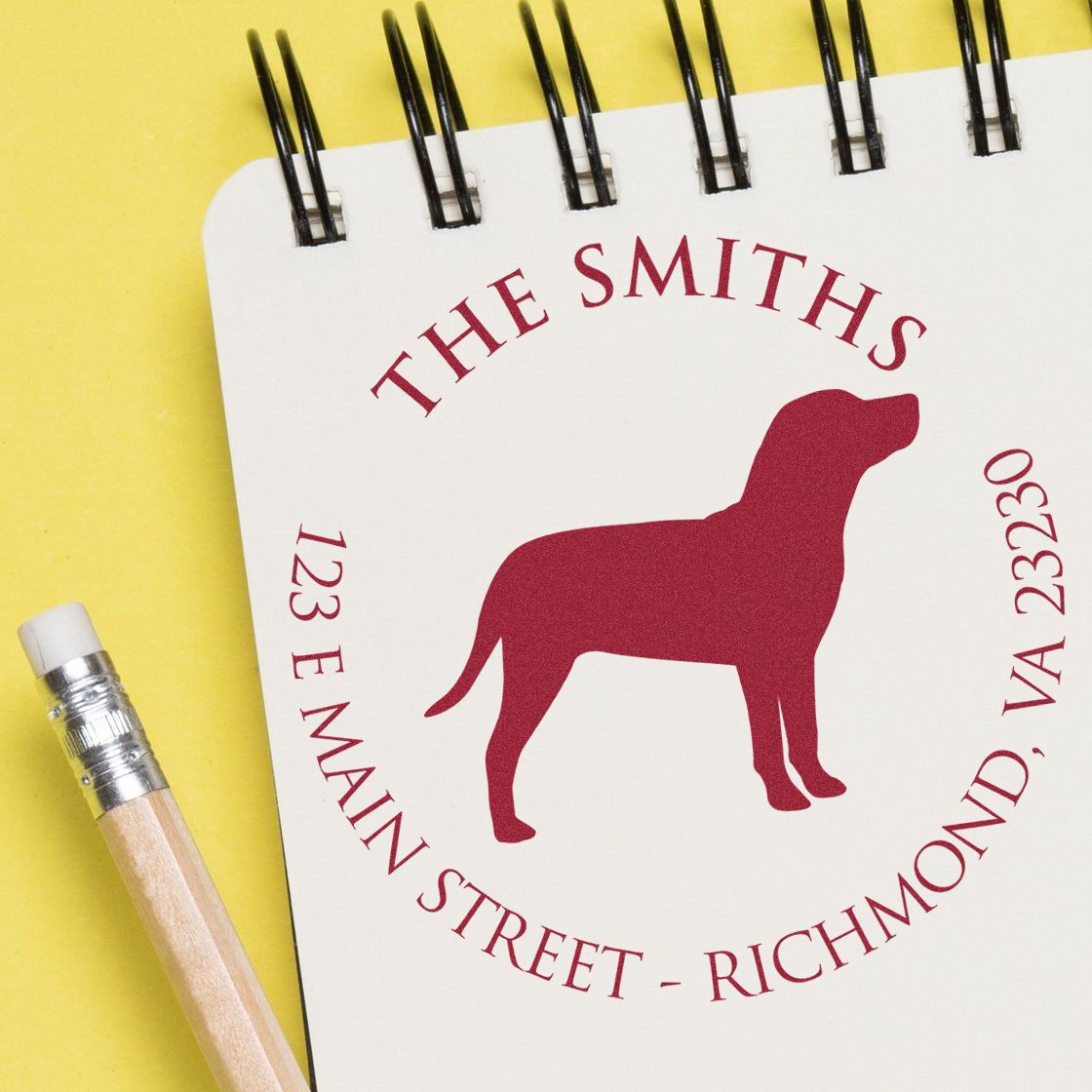 Custom dog stamp seal on a white spiral-bound notebook with maroon text, beside a pencil on a bright yellow background.