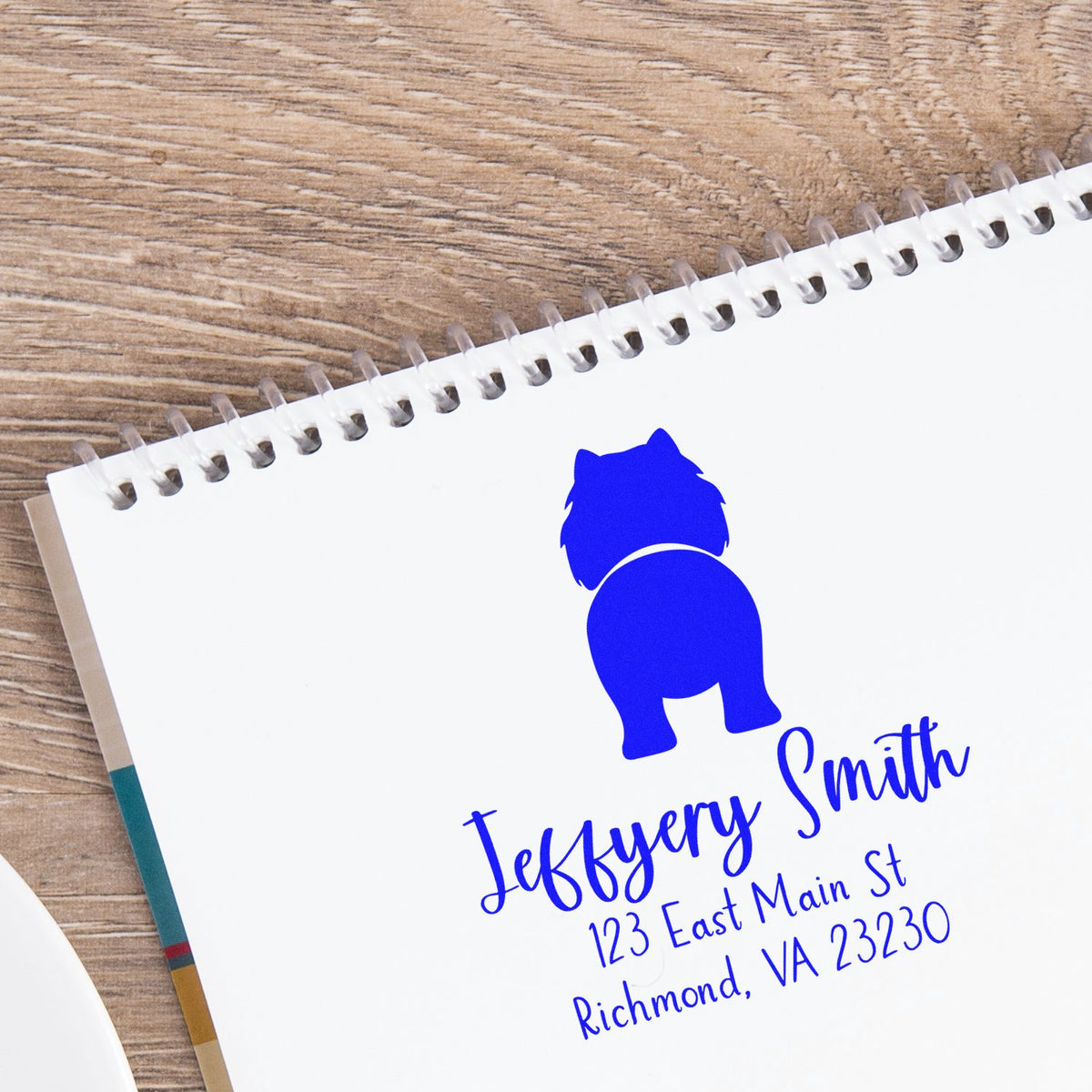 Blue custom seal stamp on white spiral notebook, with dog logo, Jefferson Smith name, and address on wood background.