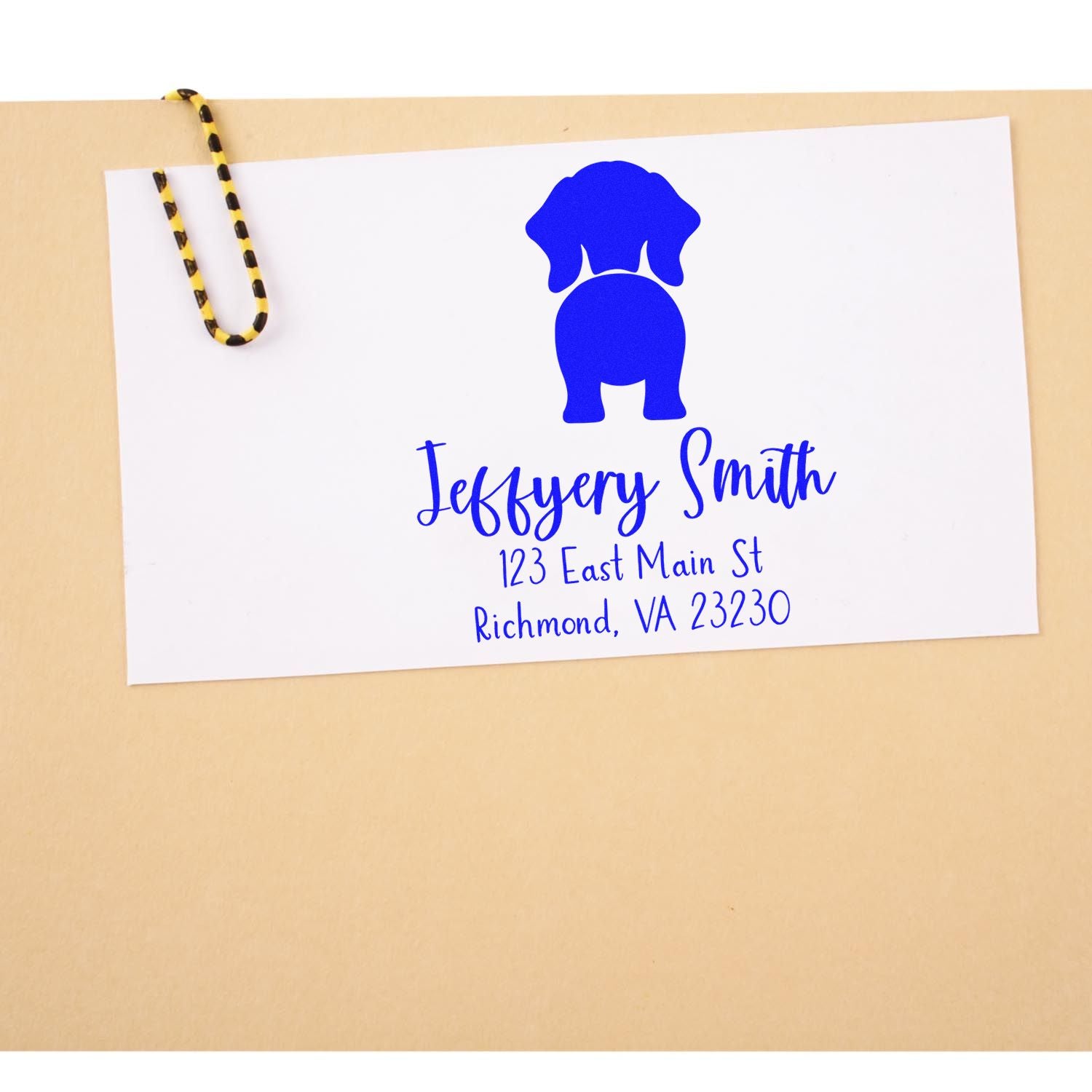 White envelope with blue custom address stamp and dog seal logo, clipped with a yellow-and-black paperclip on tan background.