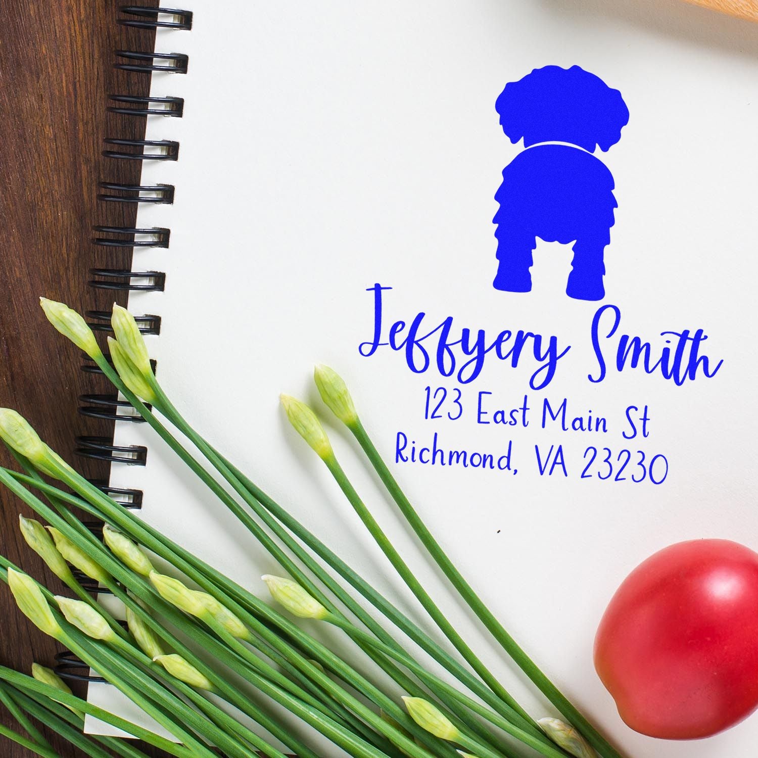 Blue personalized address stamp and seal on white notebook, with black spiral binding, green flower stems, and a red accent.