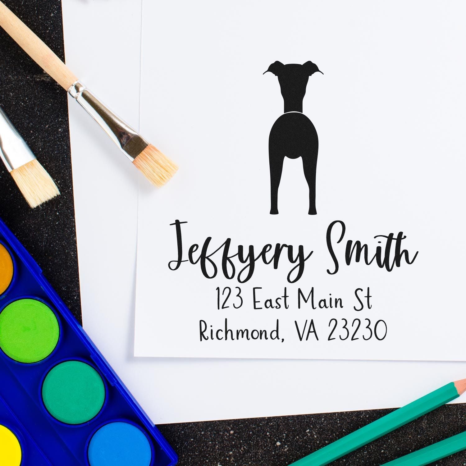 Black dog silhouette stamp and seal with Jeffrey Smith name and address, beside paint brushes and watercolor palette.