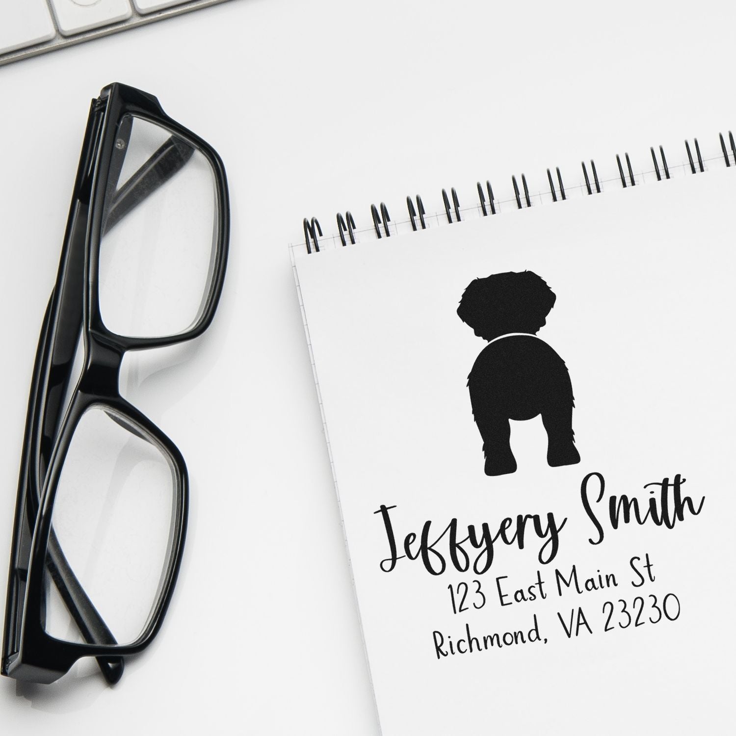 Black custom stamp and seal with dog silhouette and address on white spiral notebook, beside black eyeglasses.
