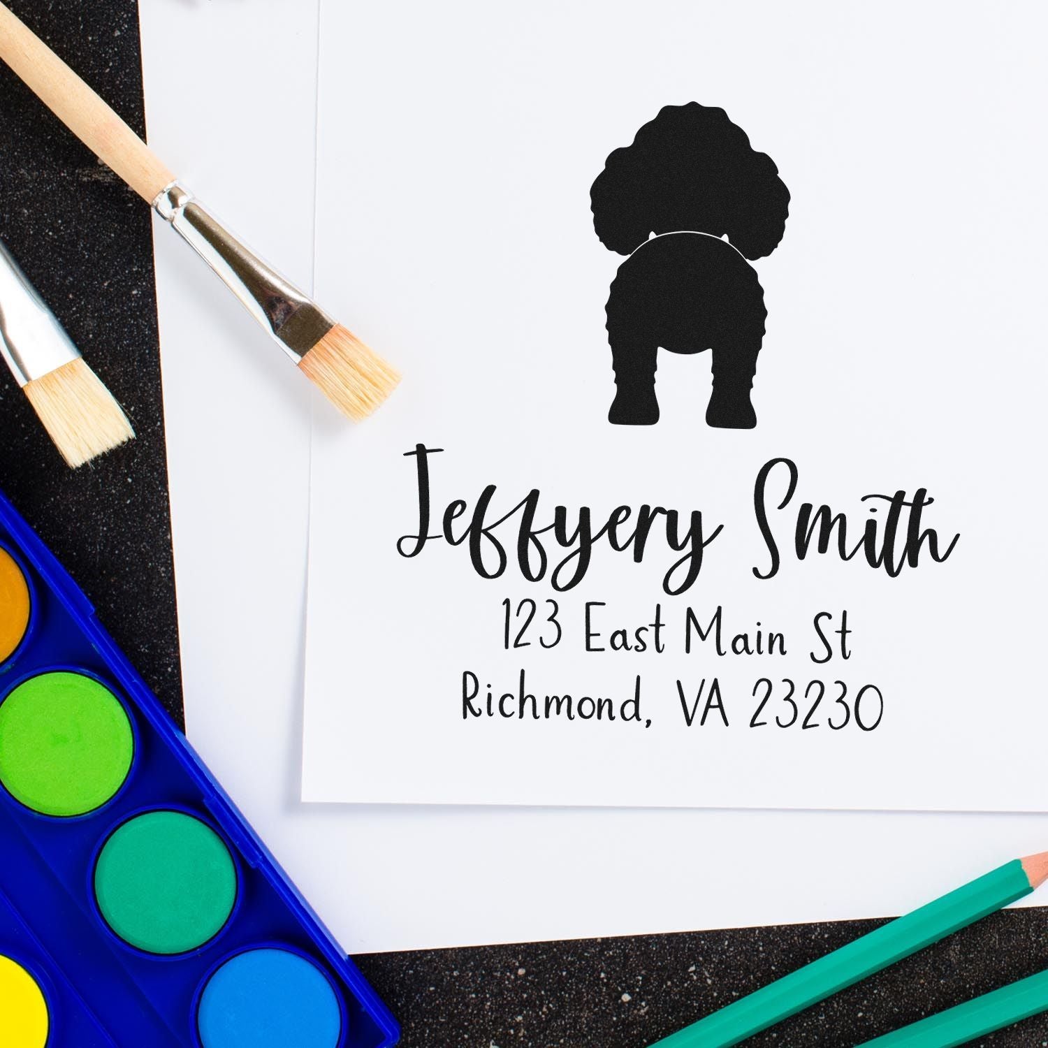 Custom black address stamp and seal with script name on white paper, framed by paintbrushes and bright art supplies.