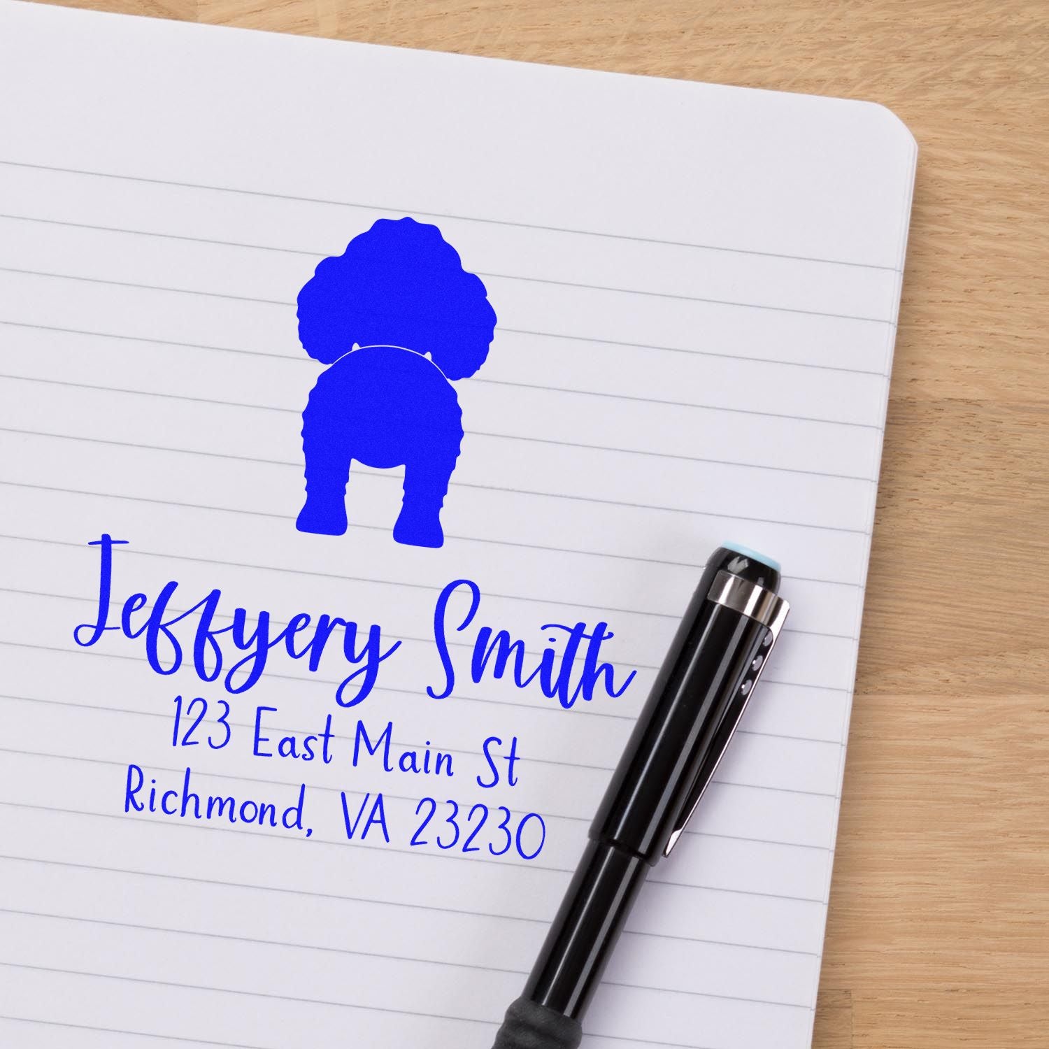 Blue custom address stamp and seal on lined notebook, with dog silhouette and black pen on a wooden desk.