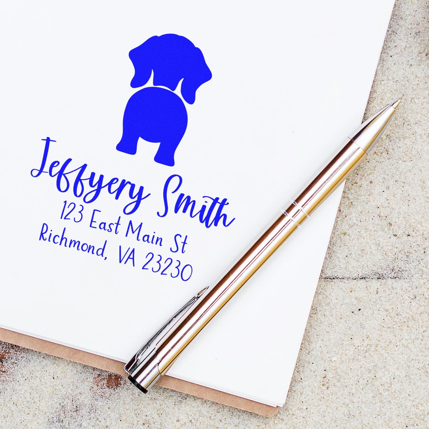 Custom blue address stamp with elephant logo and Jeffrey Smith address, beside a silver-gold pen on white paper.