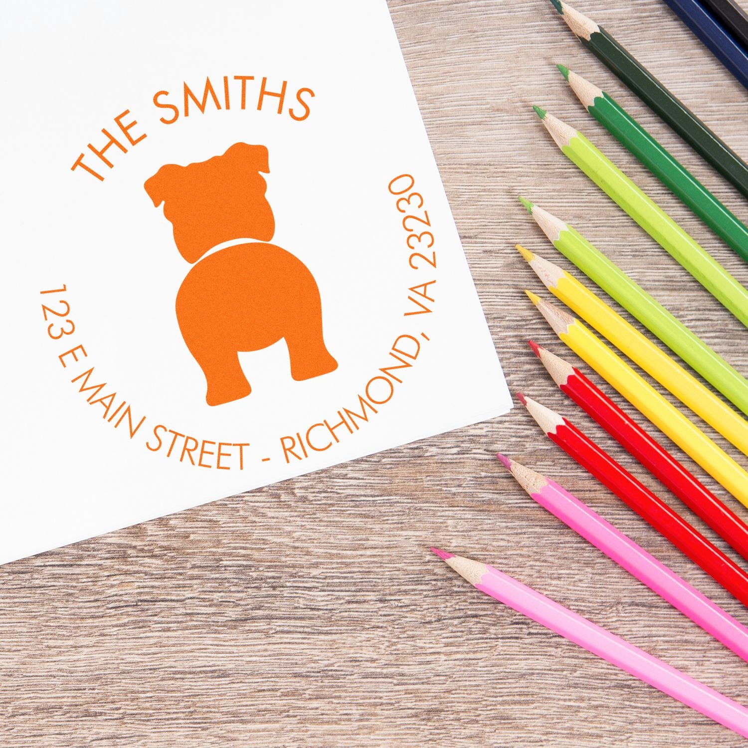 Custom orange dog address stamp seal on white paper beside colorful pencils on a wood desk.