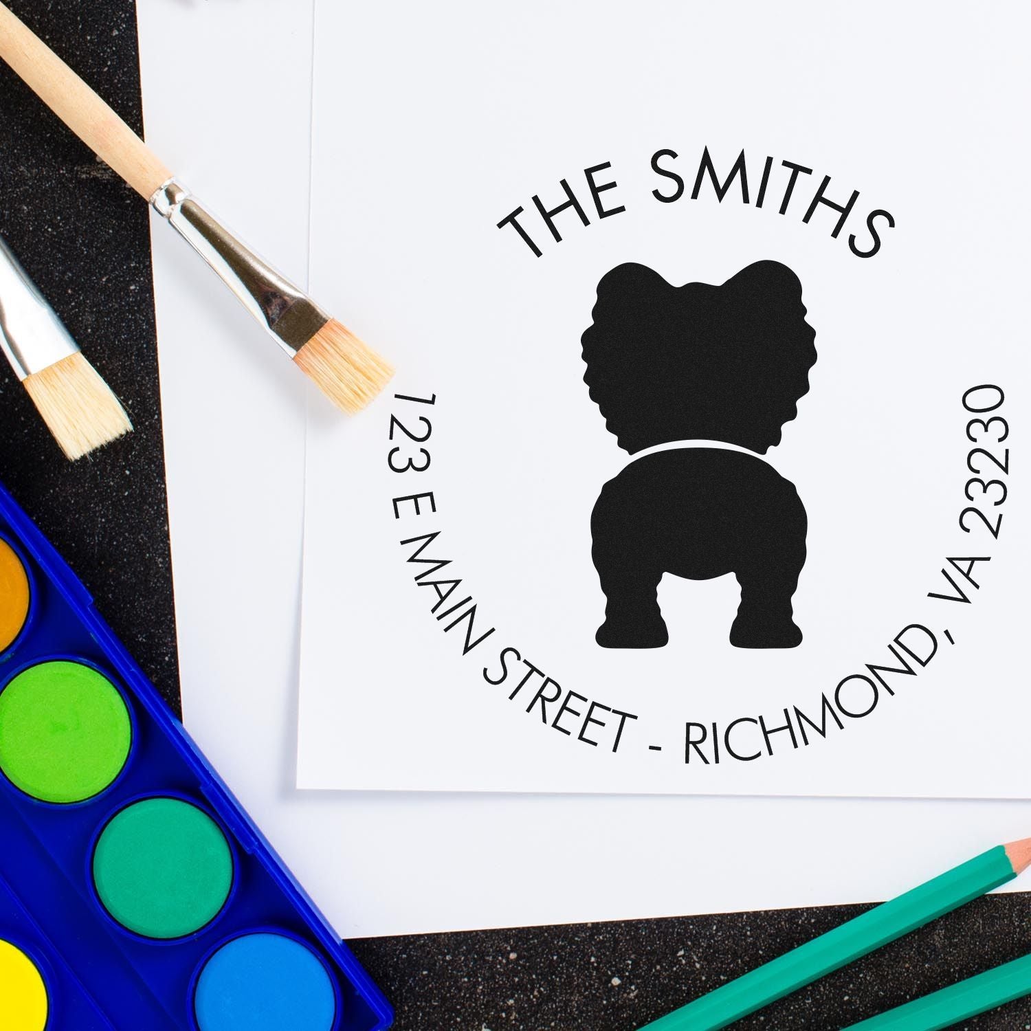 Personalized black stamp seal with bear silhouette, THE SMITHS, and 123 E Main Street, beside paintbrushes and watercolor paints.