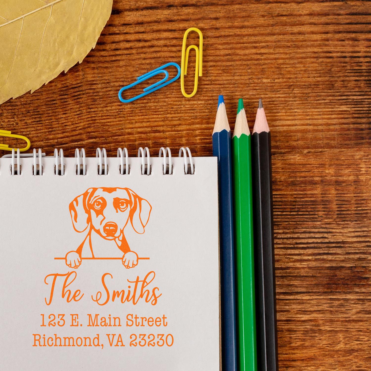 Orange custom address stamp and seal with dog illustration on white notepad, beside blue, yellow, green and black pencils on wood.