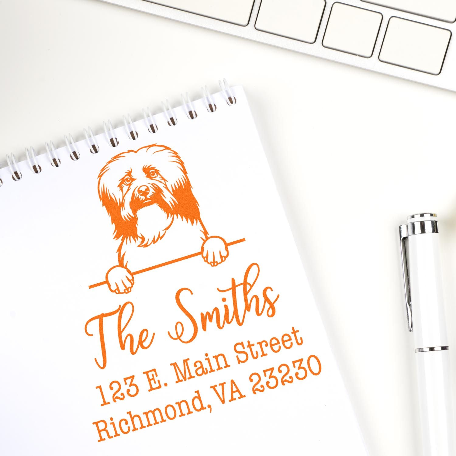 Custom orange address stamp and seal with dog logo on spiral notebook, beside white pen and keyboard