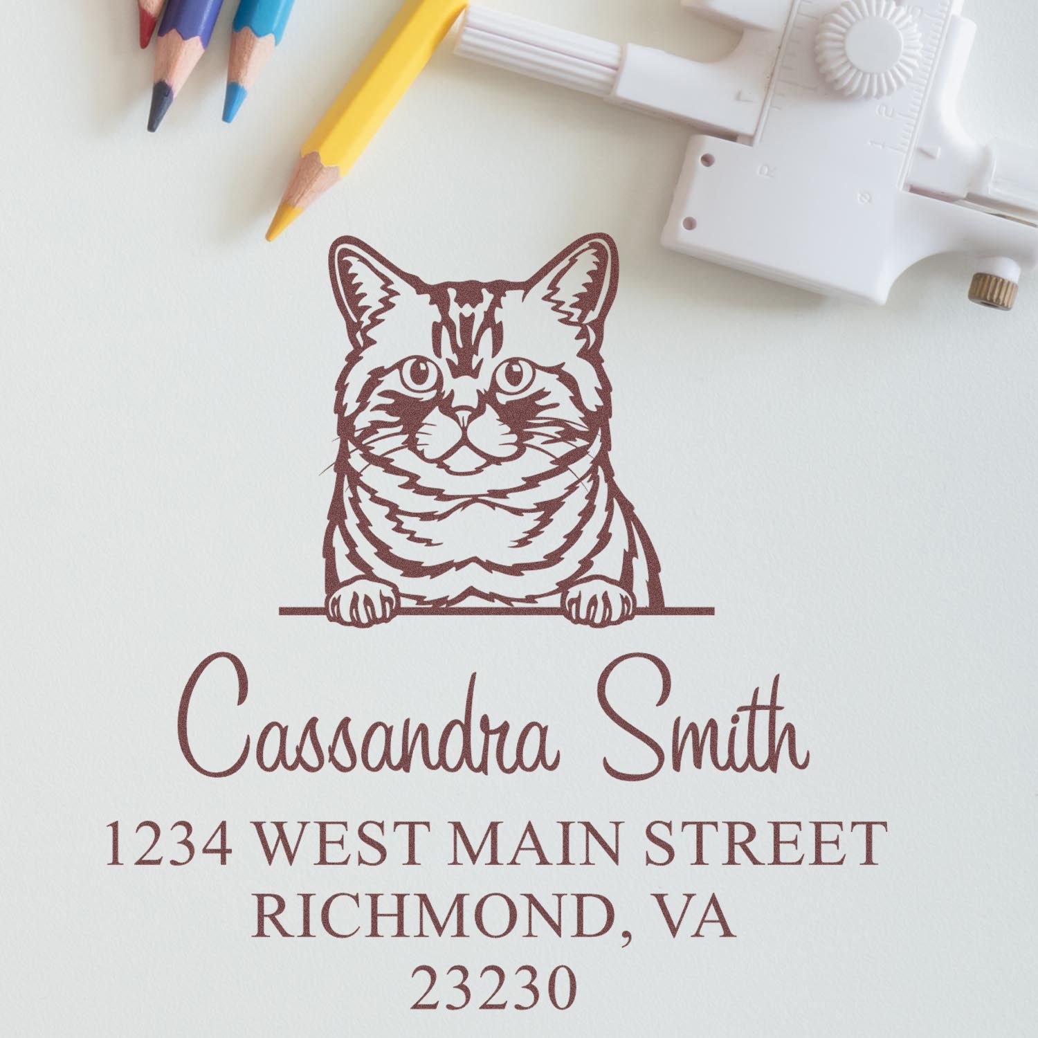 Brown custom cat return address stamp and seal on white paper, with colorful pencils and a white embossing tool.