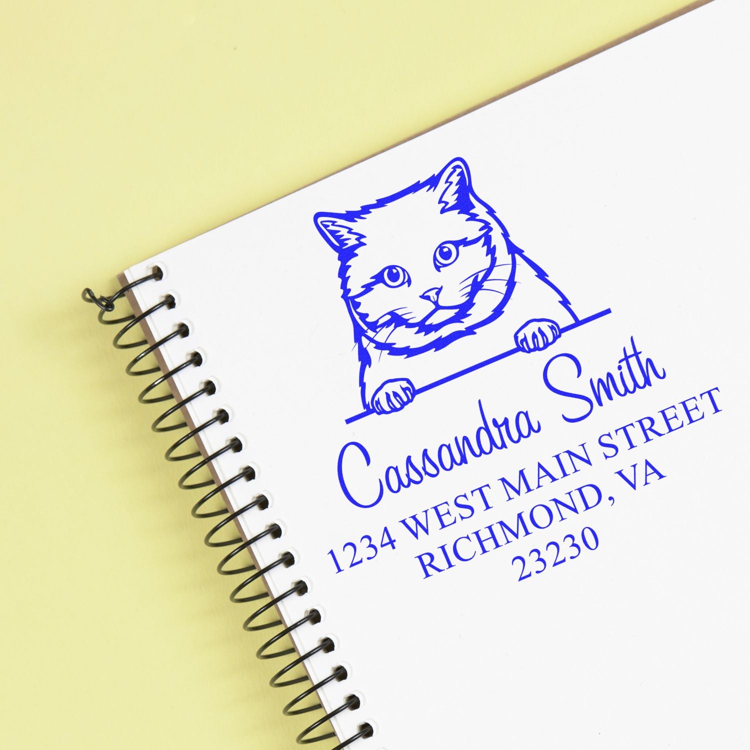 Blue cat custom address stamp in blue on a white spiral notebook, with Cassandra Smith and address on a pale yellow background.