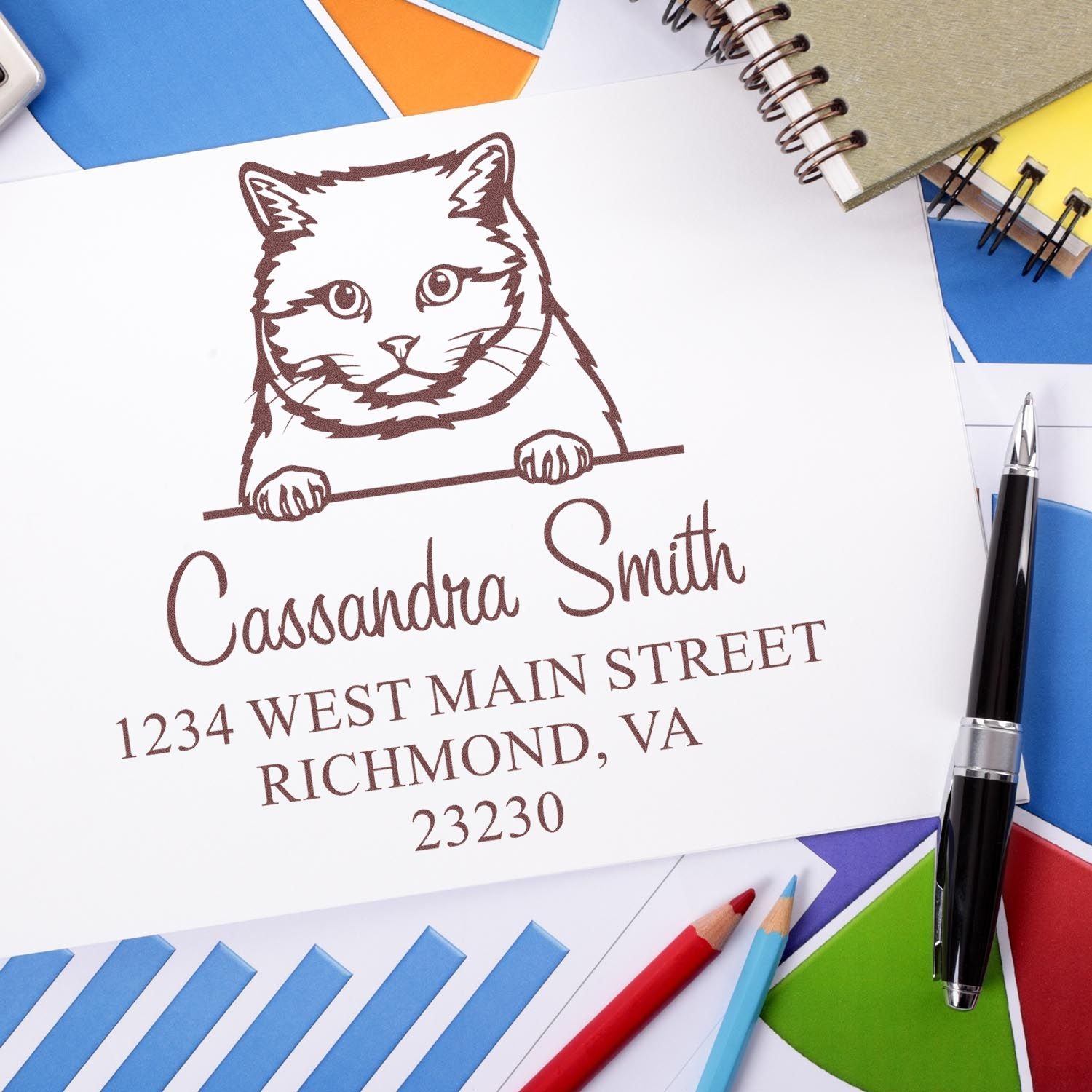 Brown custom cat stamp and return address seal on white paper, with colorful notebooks and pens.