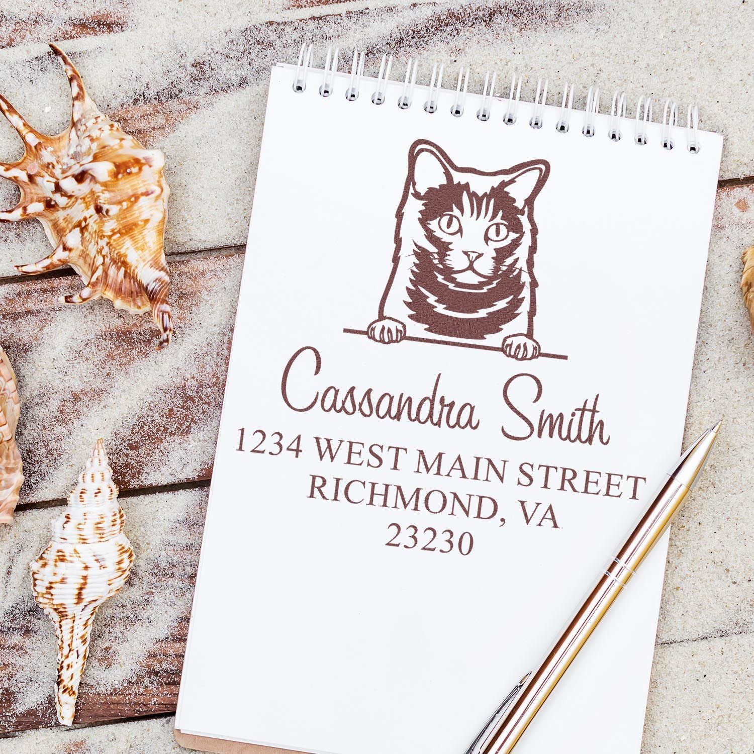 Brown cat address stamp on white spiral notebook, with custom name and address seal, seashells, and gold pen.