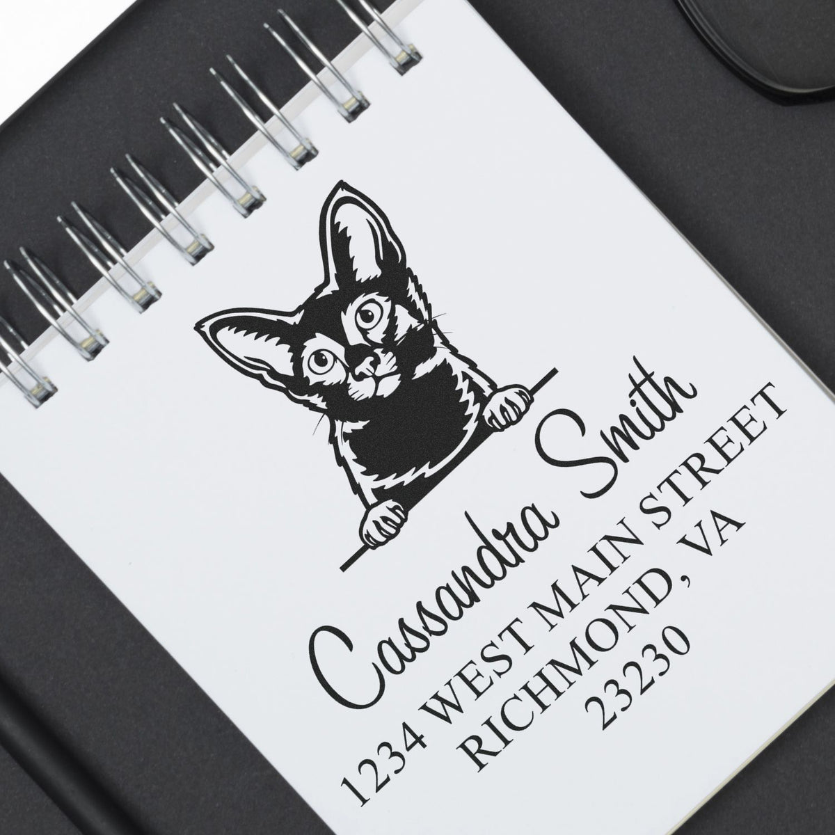 Black custom stamp and seal on a white spiral notebook, featuring a cat logo and address text.