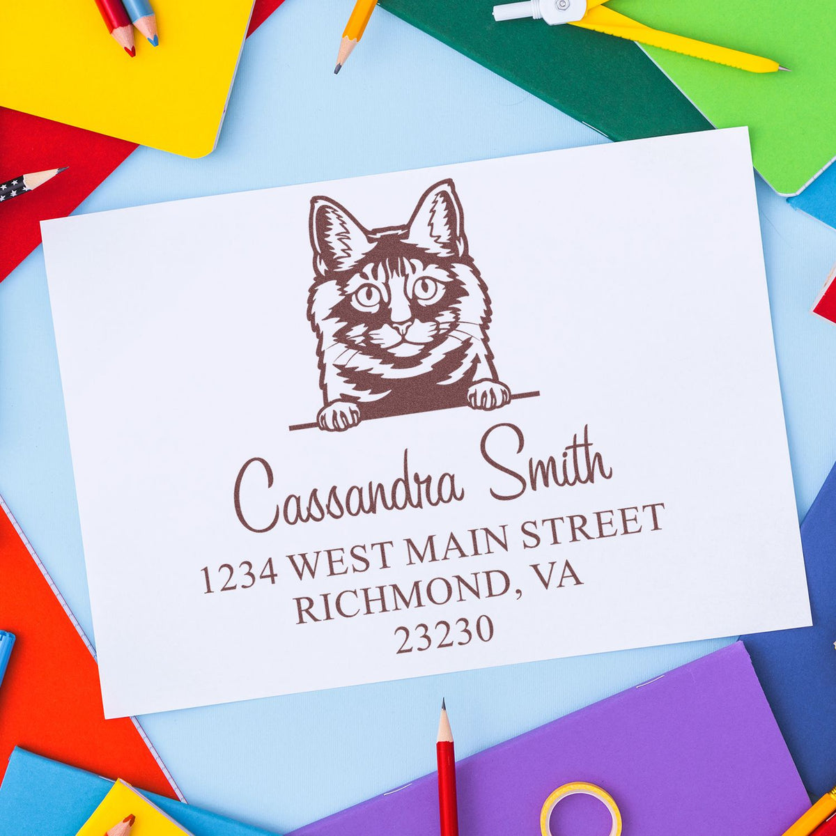 Brown cat custom address stamp and seal on white card, with colorful pencils and notebooks on a blue background.