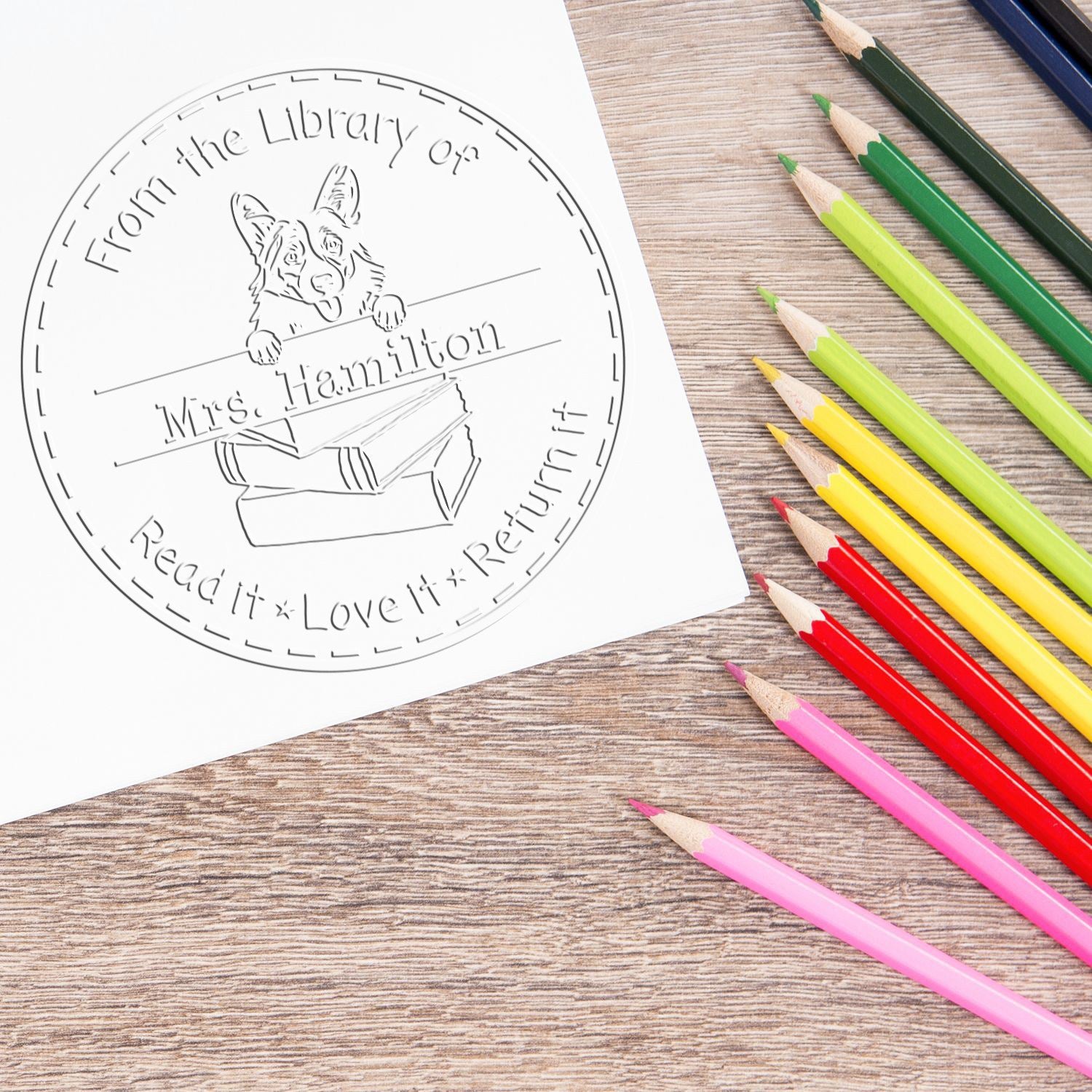 Custom stamp and seal with library dog design on white paper, beside colorful pencils on a wood desk