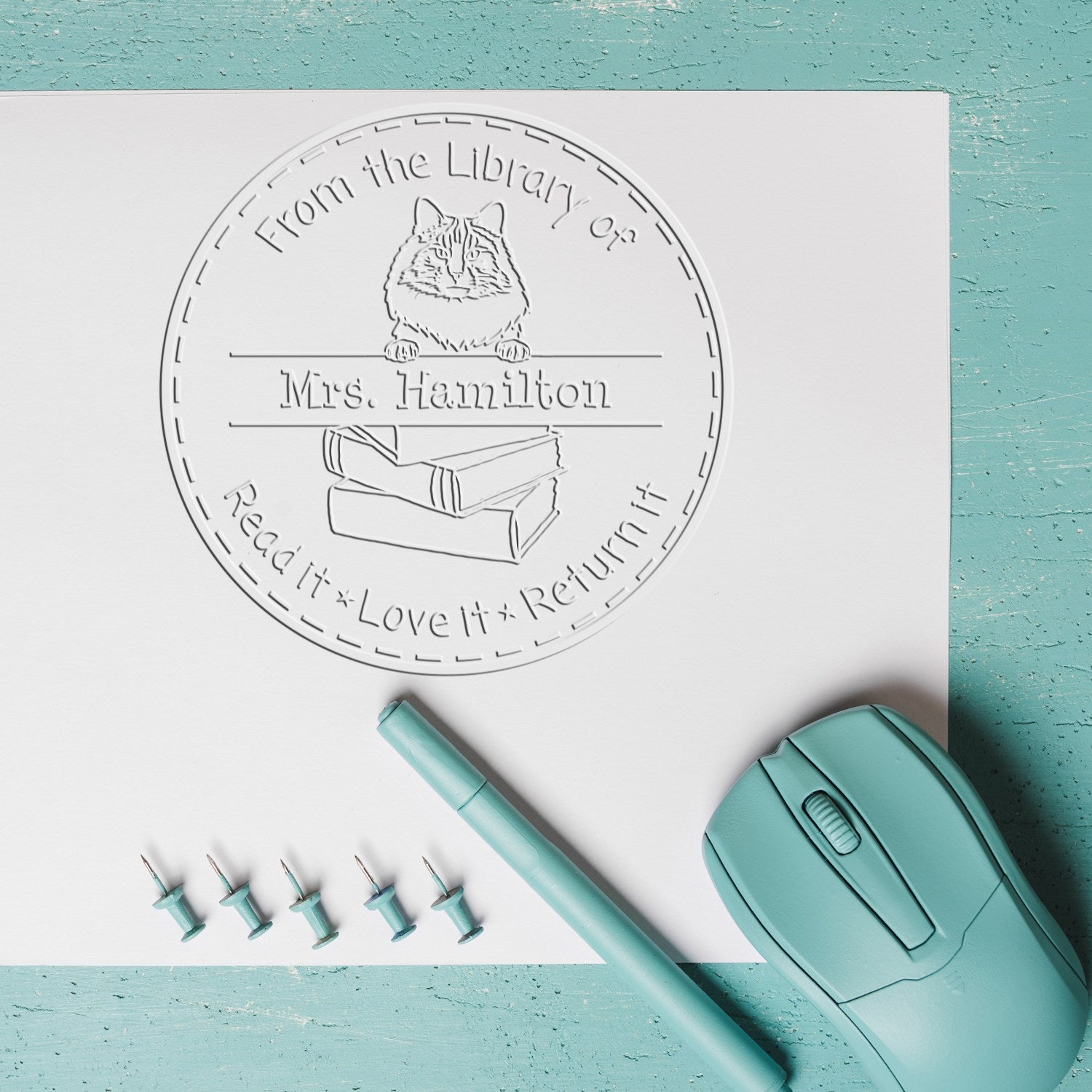 Teal custom library stamp and seal products on white paper, featuring a cat, books, and embossed Mrs. Hamilton text.