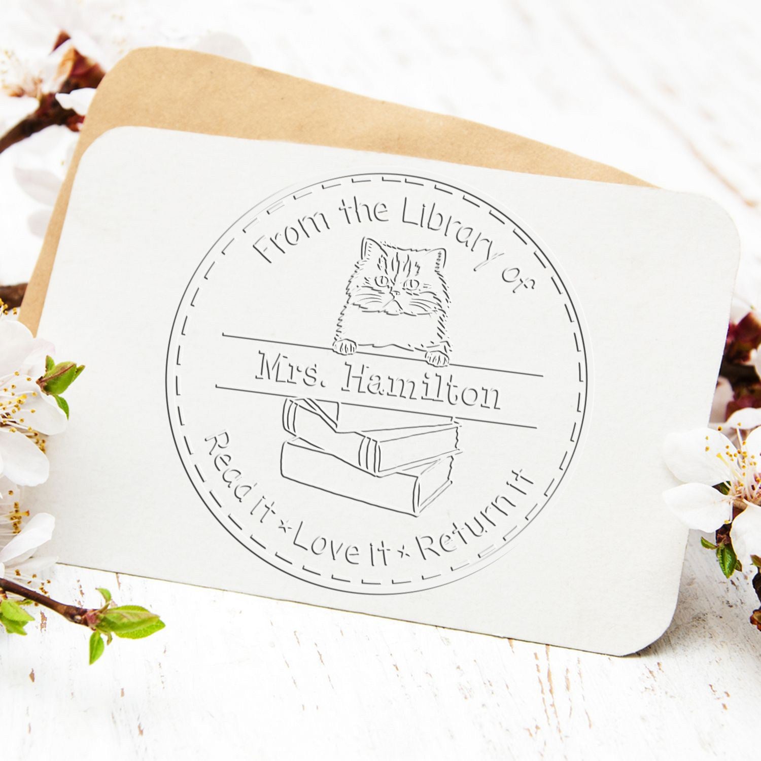 White custom library stamp seal with cat and books on a tan envelope, framed by white blossoms.