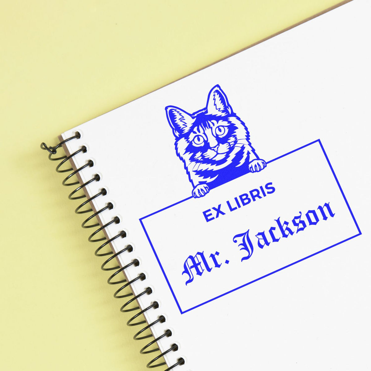 Blue ex libris stamp and seal with cat design and Mr. Jackson text on a white spiral notebook, pale yellow background