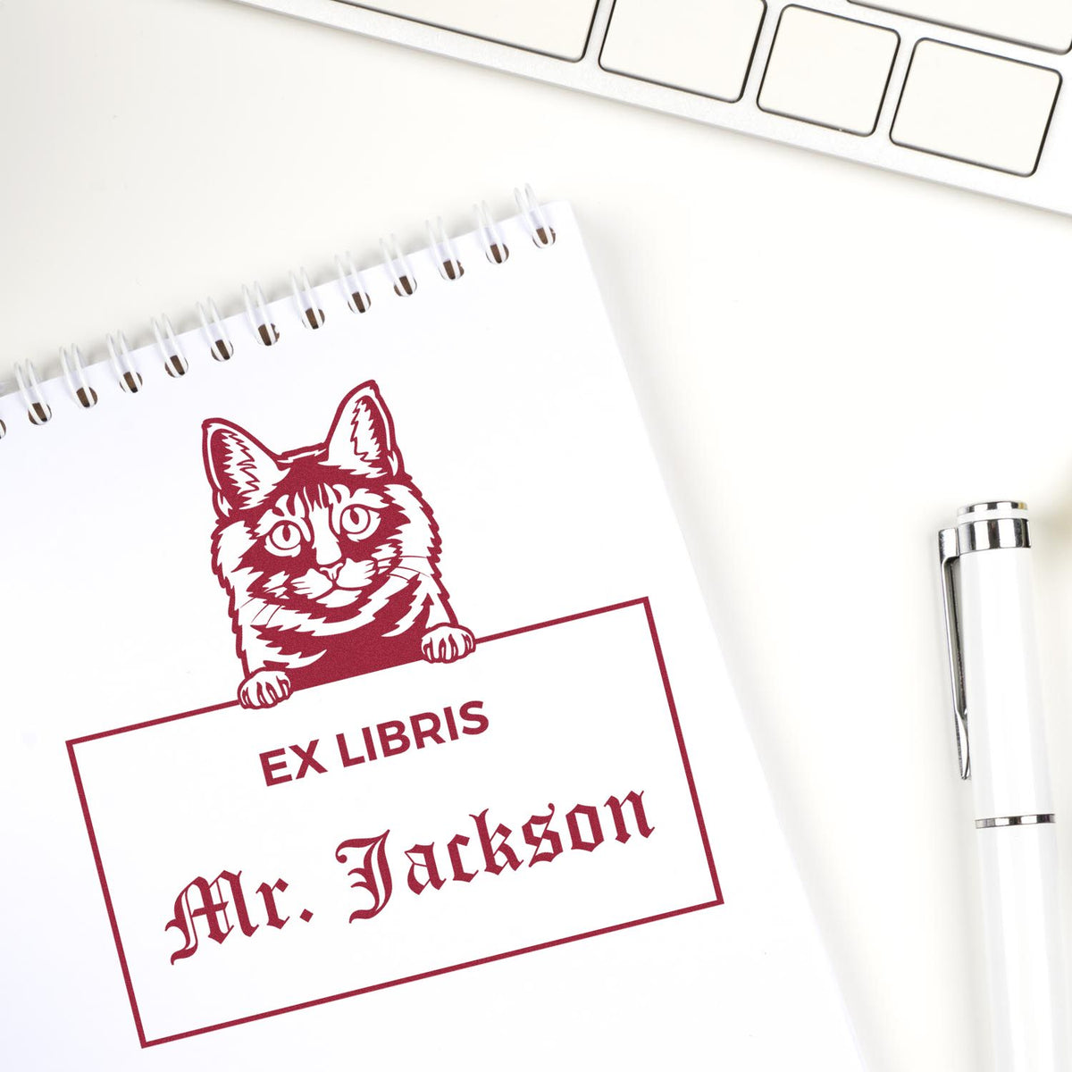 Burgundy custom ex libris stamp seal on white spiral notebook with cat and Mr. Jackson, beside a white pen and keyboard.