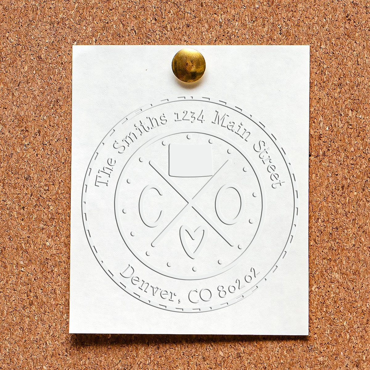 White embossed custom circular stamp seal pinned to corkboard with brass tack, featuring name text and Denver, CO.