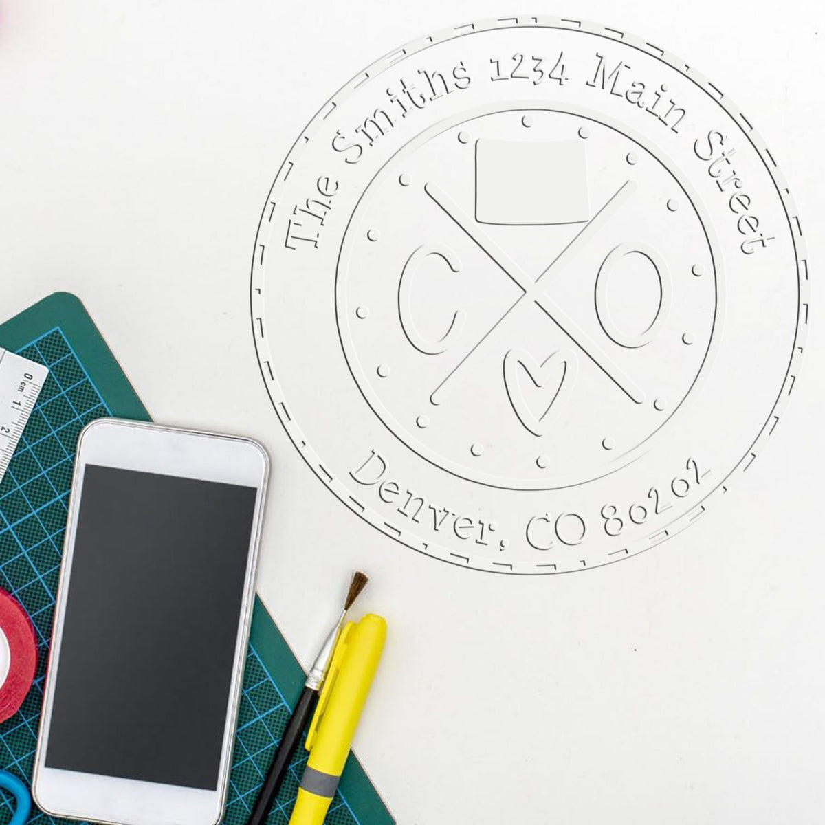 White round custom seal stamp with embossed text and logo, beside green cutting mat, smartphone, brush, and yellow cutter.