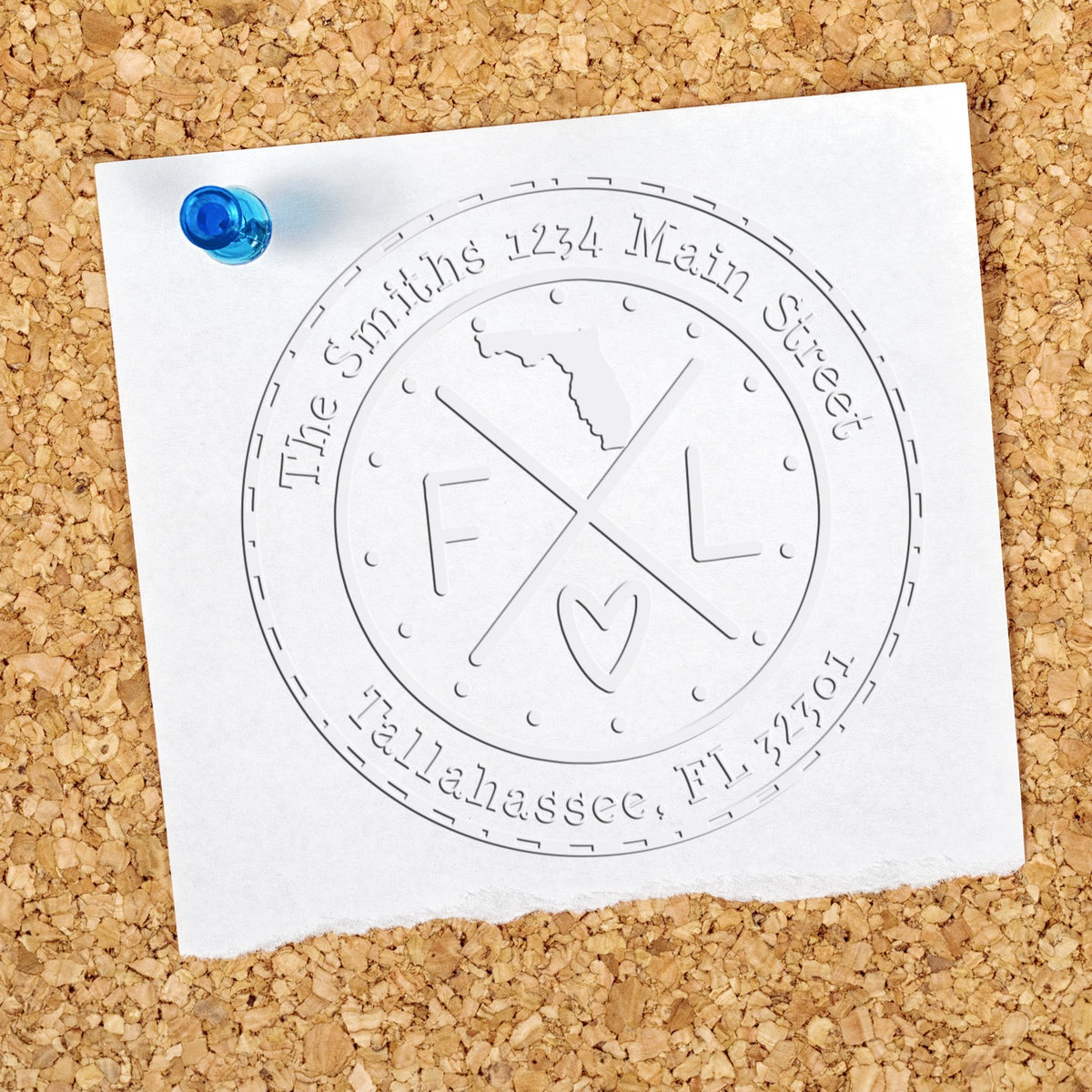 White embossed circular seal stamp on pinned paper with blue pushpin, on tan corkboard.