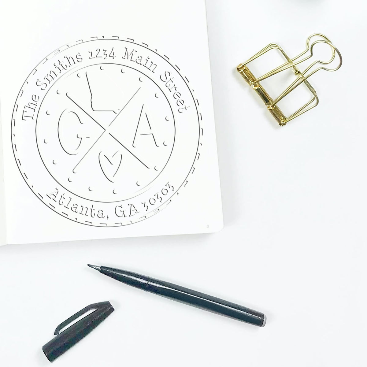 Custom stamp and seal products: circular monogram design on white paper with gold binder clip, black pen, and cap.