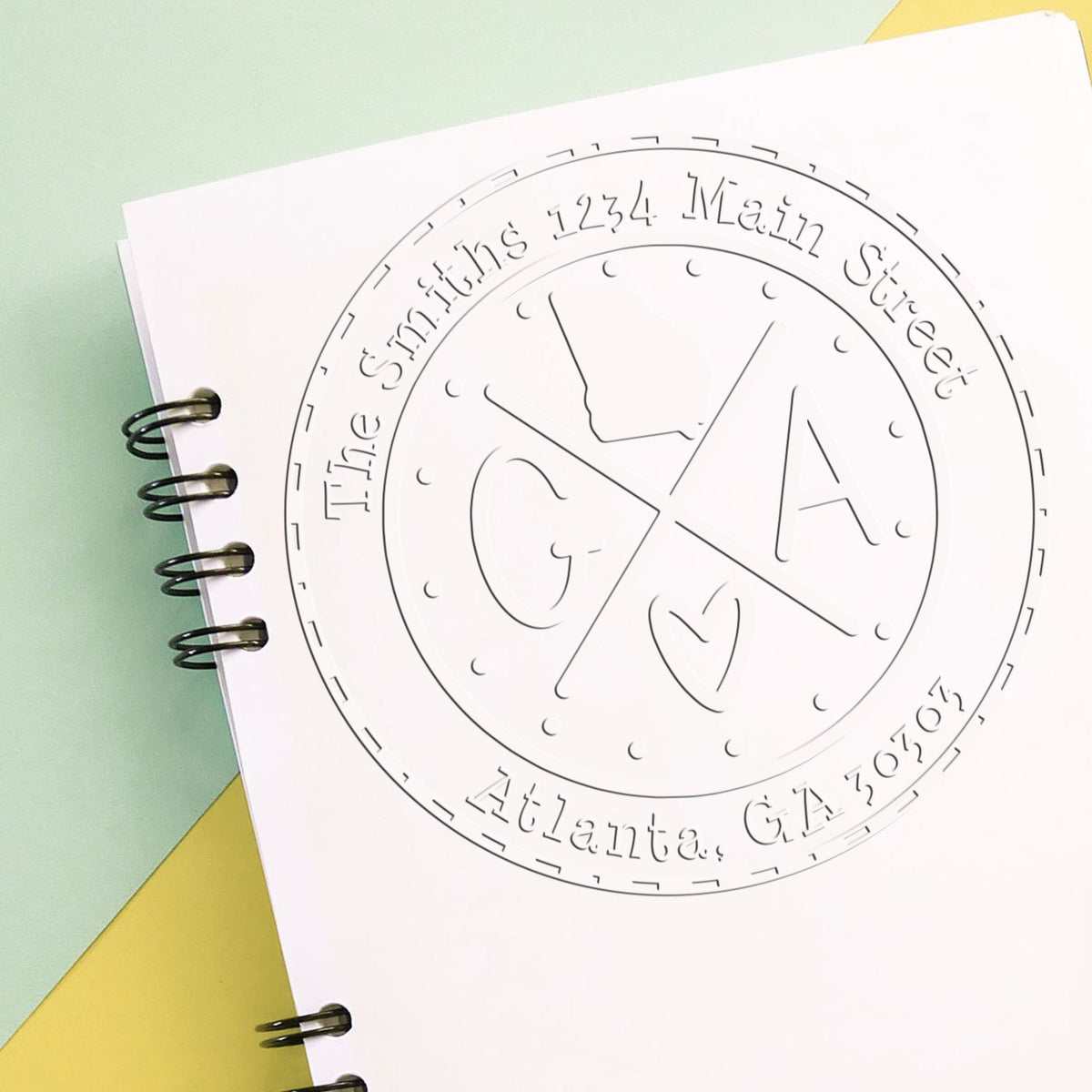Custom circular stamp seal on white spiral notebook, black line design with GA and heart logo, pastel green and yellow background.