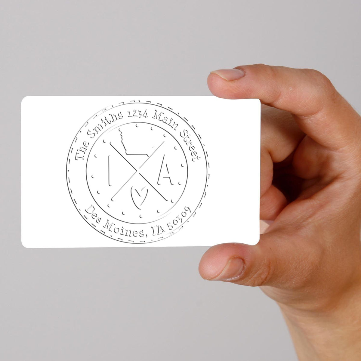 Hand holding white stamp/seal card with gray circular seal reading The Smiths, 1234 Main Street, Des Moines, IA 50209.