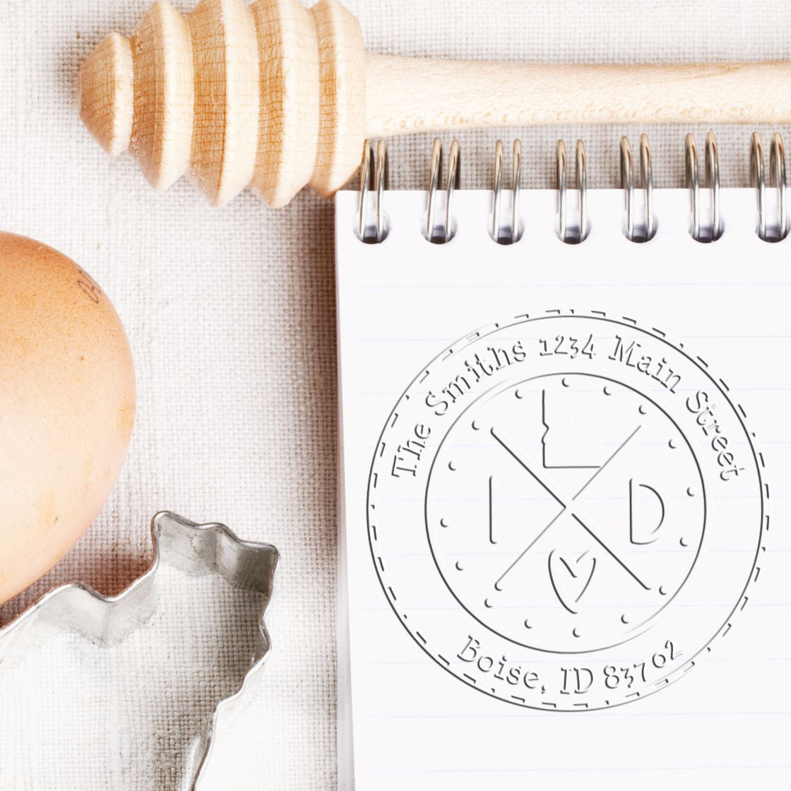 Wooden rubber stamp and circular seal impression on spiral notepad, with beige egg and metal cutter on a textured linen background.