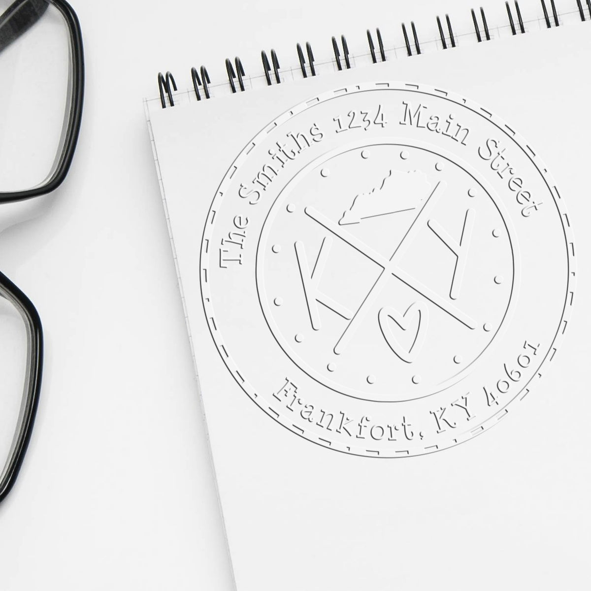 White notebook with embossed circular stamp seal reading The Smith's 1234 Main Street, Franklin KY 40601; black glasses edge.