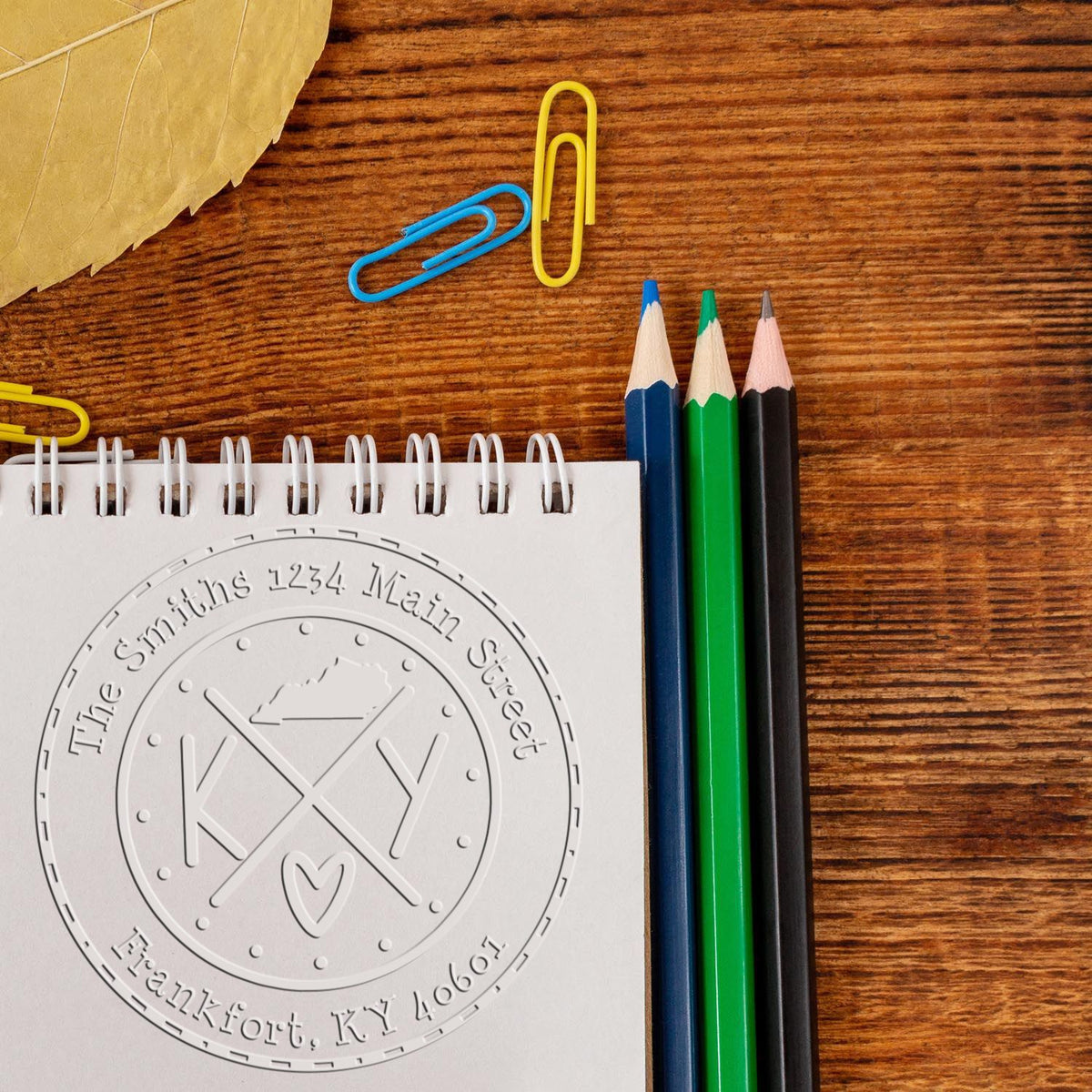 Custom embossed stamp and seal on white notepad beside blue, green, and black pencils, with yellow and blue paper clips on wood.