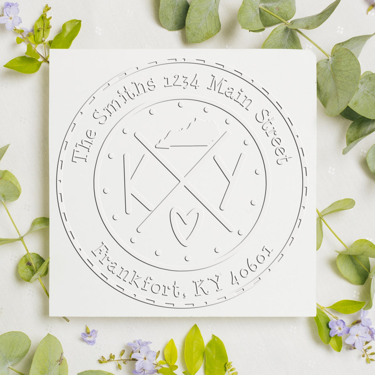 White embossed custom seal stamp with KY monogram and address, surrounded by green leaves and purple flowers on a white background.