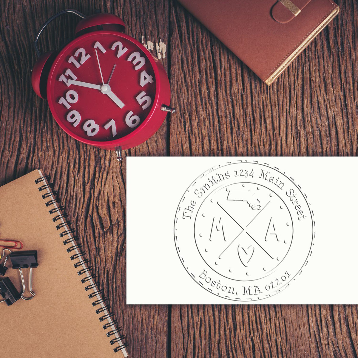 Red clock on wood desk beside notebook, leather folder, and custom round stamp and seal for The Smiths, Boston, MA.