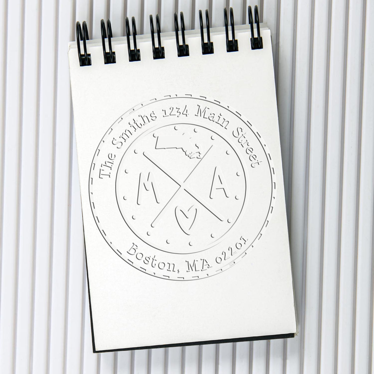 White spiral notebook showing a custom round embossing stamp seal, black rings, and engraved Boston, MA text.
