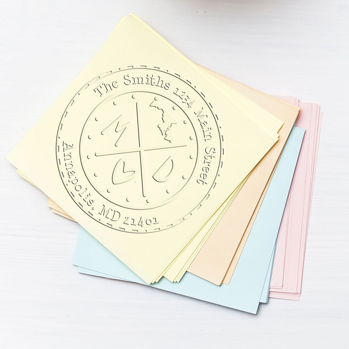 Custom embossing seal on stacked pastel paper sheets in cream, blue, pink and beige, with a circular family name and address design.