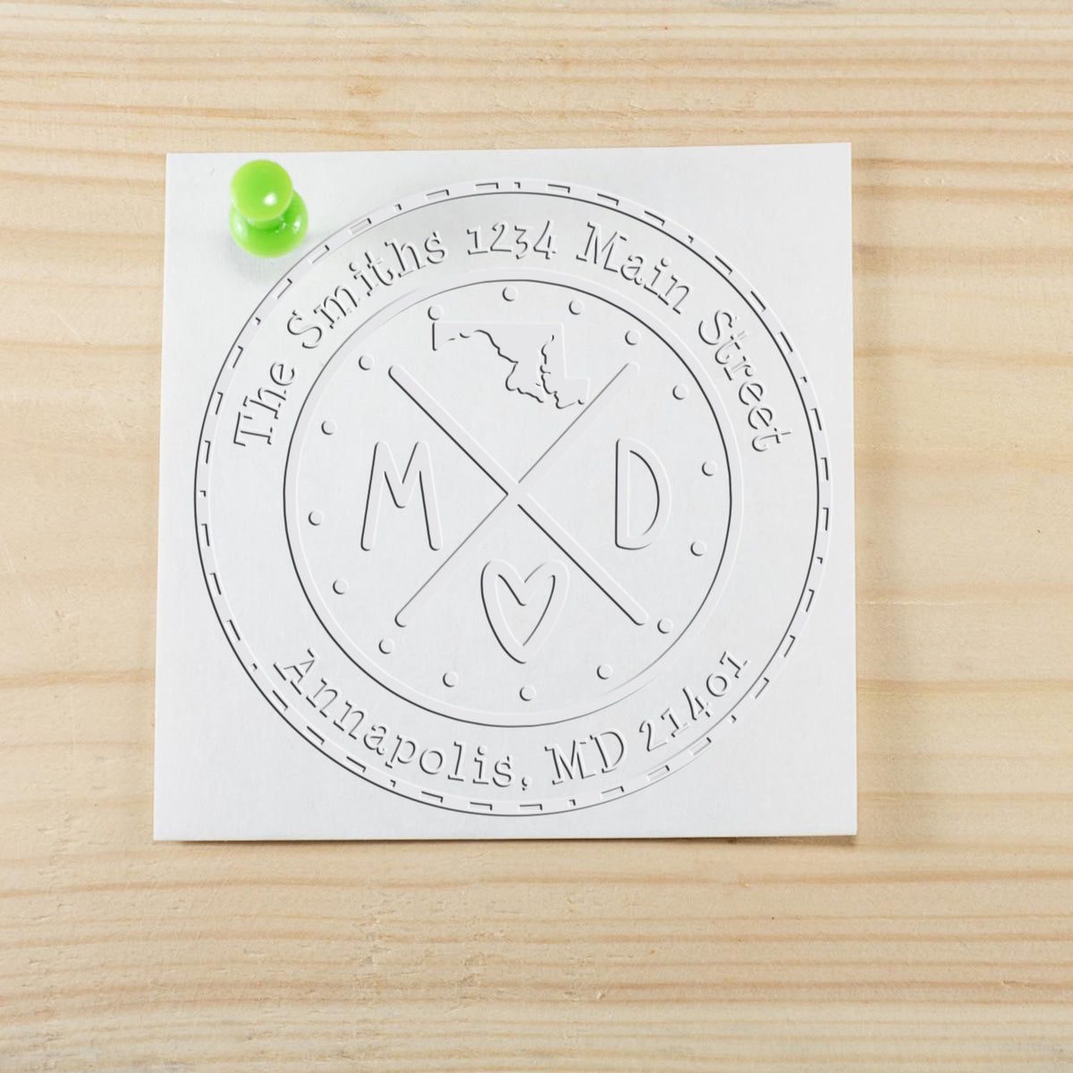 White embossed custom stamp seal with MD initials and circular border, pinned with a bright green tack on light wood.