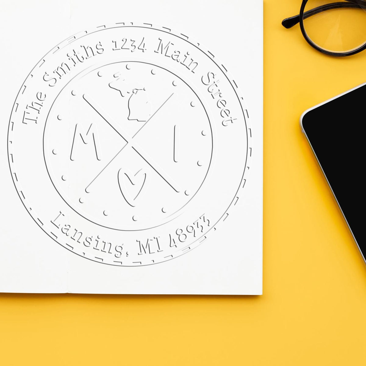 Custom circular seal stamp embossed on white paper, with gray lettering on a yellow desk beside black glasses and a smartphone.