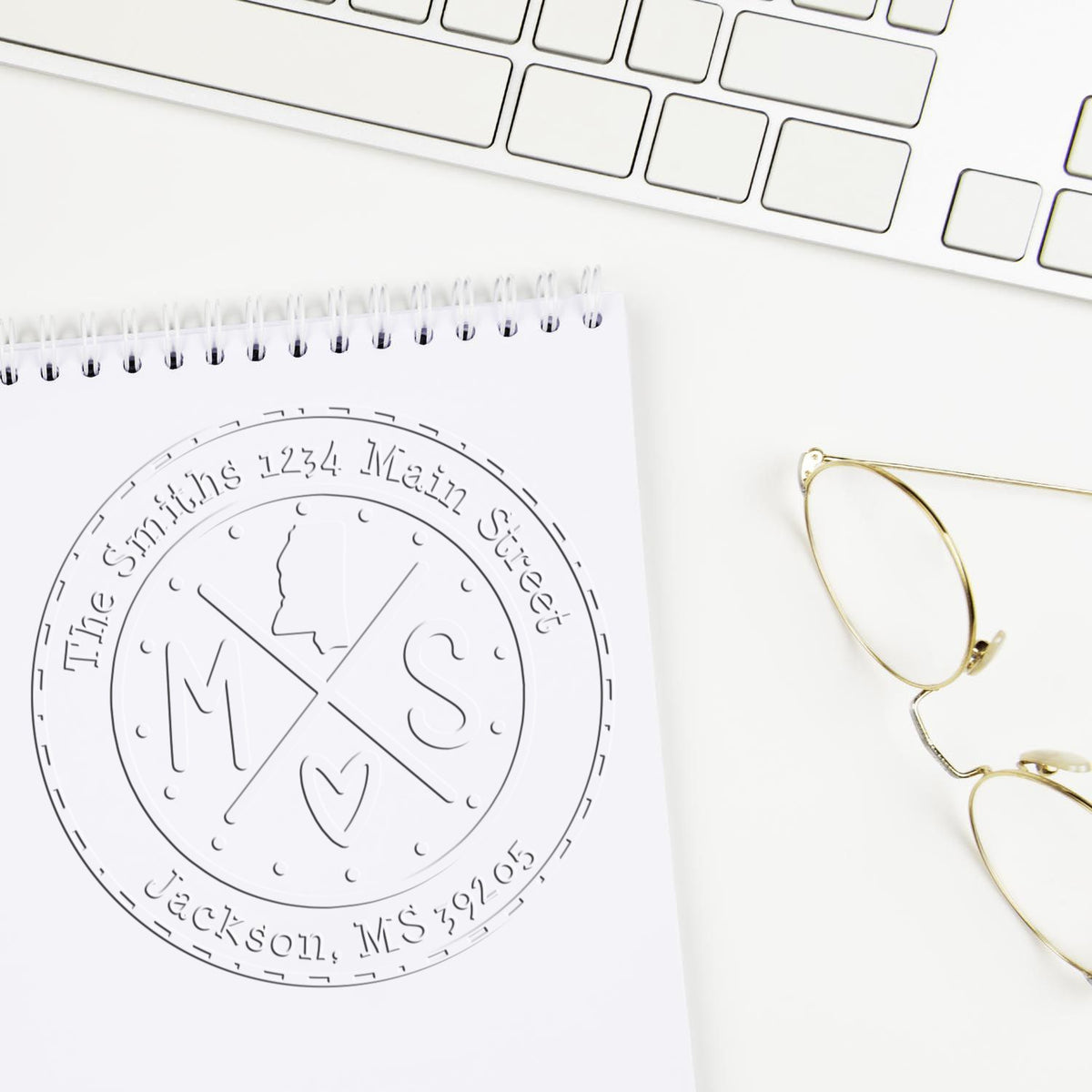 Custom embossed stamp seal with circular monogram on a white notepad, beside a keyboard and gold eyeglasses.