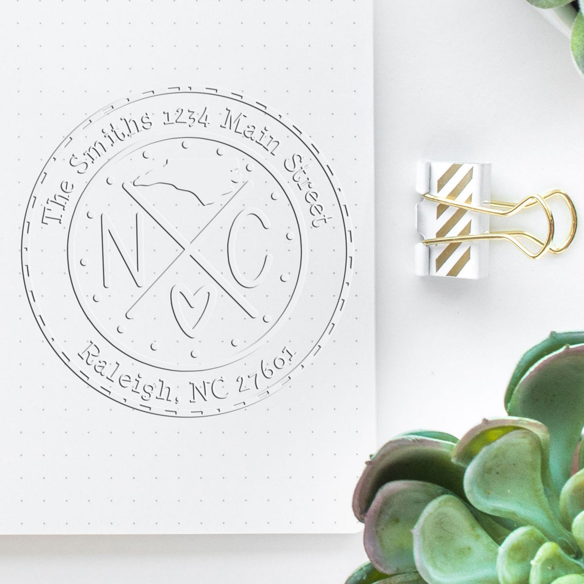 White notebook page with embossed circular NC seal stamp, gray text, gold paper clip, and green succulent accents.