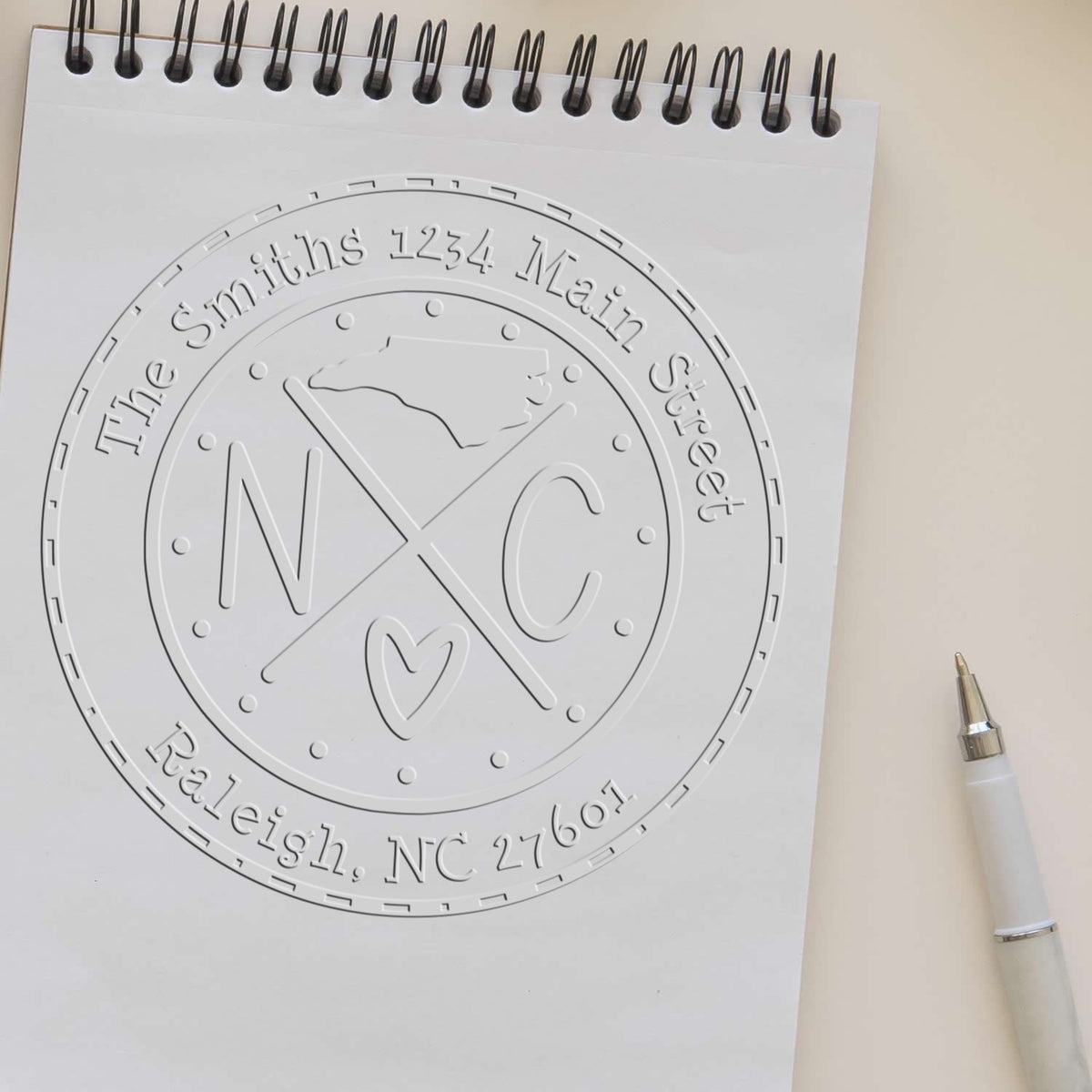 White embossed custom stamp and seal proof with circular monogram, address, and heart on a spiral notebook beside a white pen.