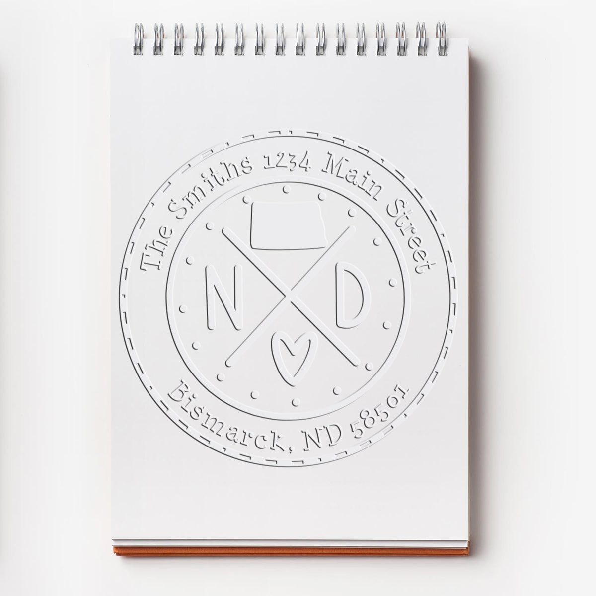 White spiral notebook with an embossed custom seal stamp, circular logo, crossed initials, heart, and address text.