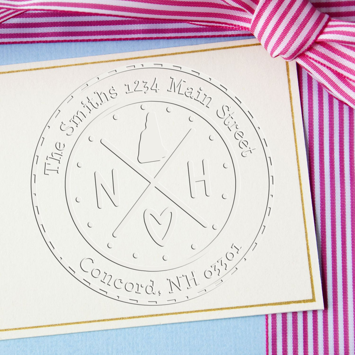 White embossed monogram stamp seal on cream card with gold border, pink striped ribbon, and blue background