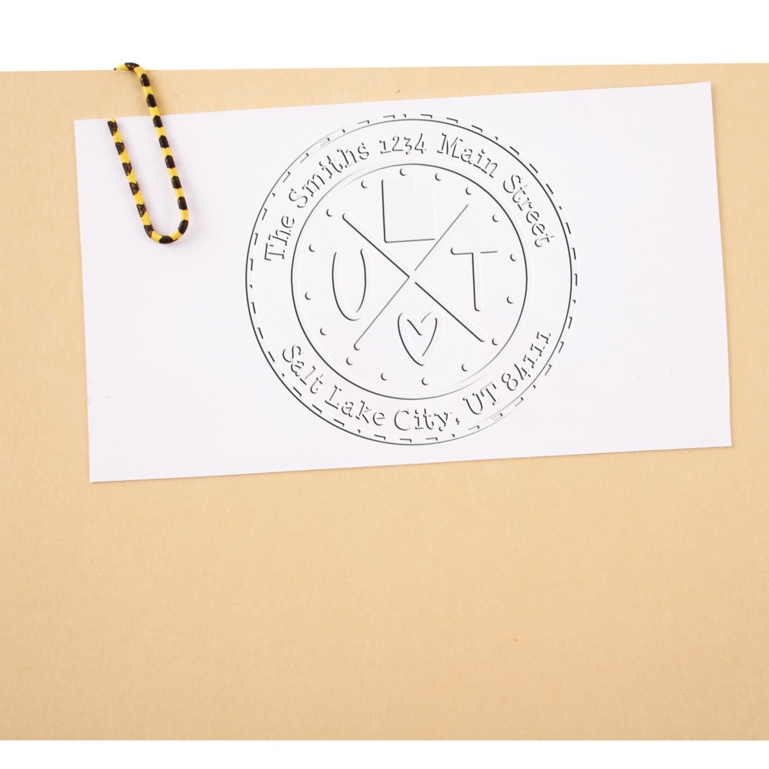 White sample stamp seal on paper with circular embossed logo, black text, clipped with yellow-and-black cord on tan background.