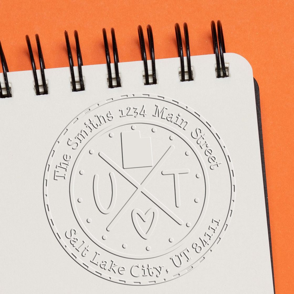 Embossed white custom seal stamp with circular family address design on a spiral notebook, against an orange background.
