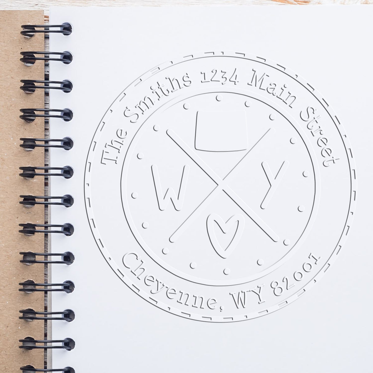 Embossed round white stamp seal on a spiral notebook, with W and Y initials, a heart, and black binding rings.